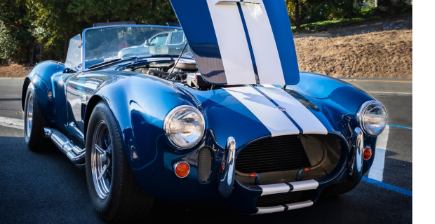 A gorgeous 1967 Ford Shelby Cobra 427 sits on a road. The metallic sparkling blue car with white stripes on the hood has the hood open, the hood opens from the base by the windshield. A truly magnificent collector car.
