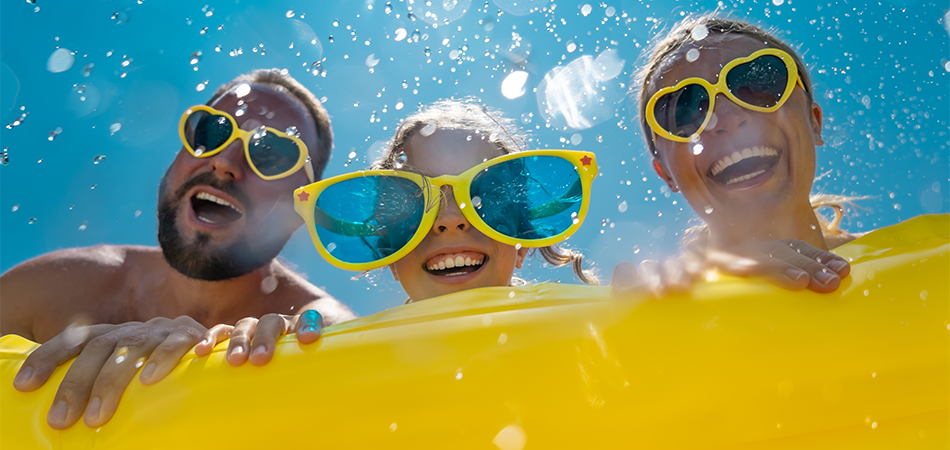 Fun summer scene with three people behind a yellow pool float, wearing colorful sunglasses and goggles, with water splashes and a bright blue sky.