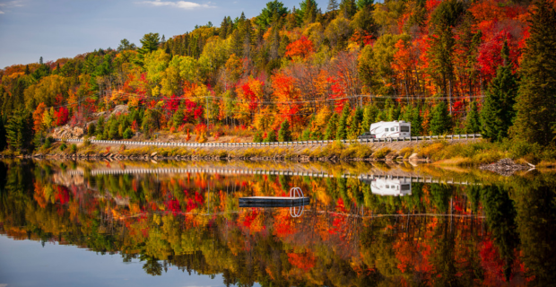 A tent trailer is driving near a fall forest with colorful autumn leaves and highway 60 reflecting in Lake of Two Rivers. Algonquin Park, Ontario, Canada.