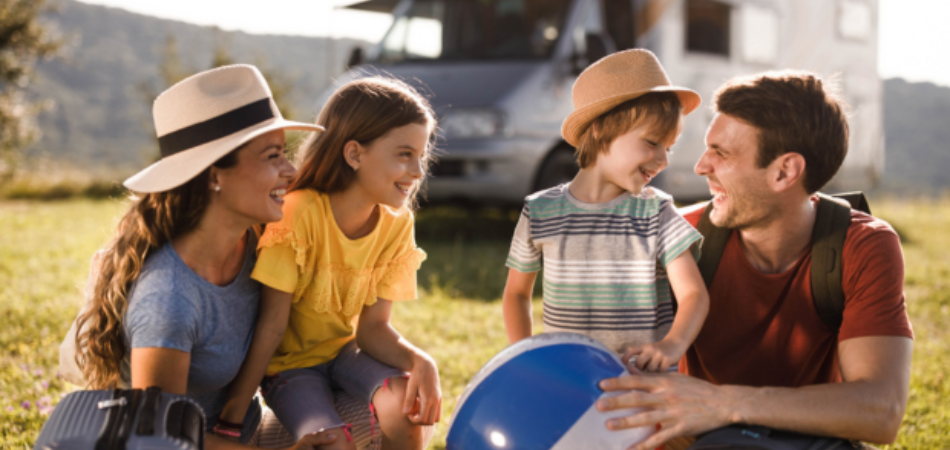 A family of four sits on the grass in front of a motorhome, dressed in casual summer clothes; one child is holding a beach ball.