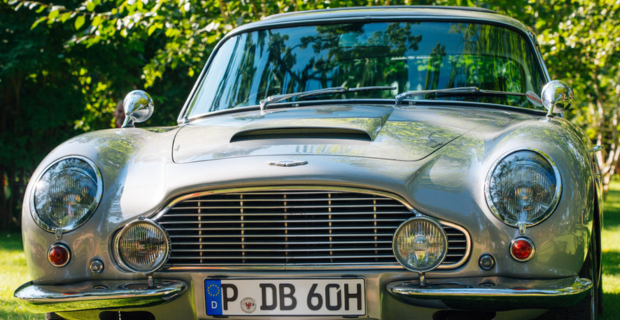 A close up of the grill and front of the stunning DB5. The steel grey paint is pristine and reflects the sun. In the background green leaves of trees can be seen