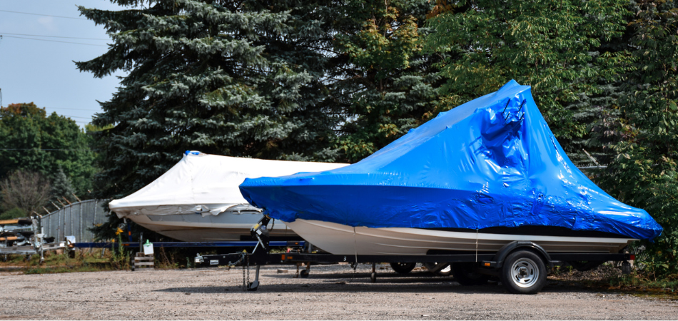 Two boats sitting in a winter boat storage lot next to ever green trees. One has a white cover and the other has a blue cover. 