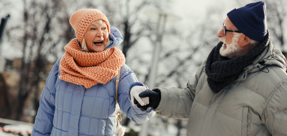 Two people dressed in heavy winter coats, scarves, and gloves walking hand in hand on a snowy path, illustrating a winter safety guide.
