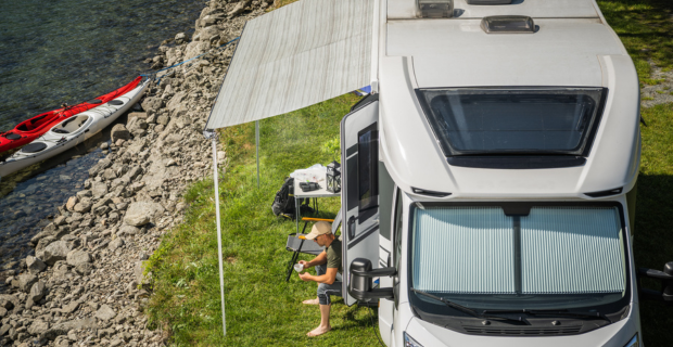 A modern class c motorhome parked next to a lake with the awning out, the driver is sitting underneath having a coffee.  