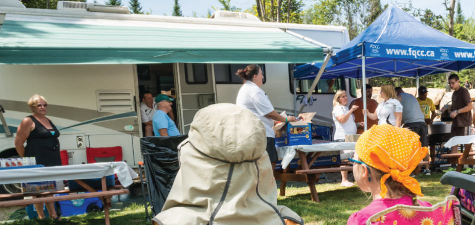 FQCC: People gather outdoors beside an RV and tents during a community event with tables and supplies set up.