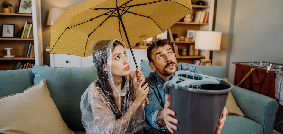 A man sitting next to his partner on their couch in their home holding a bucket to catch water leaking from the roof while his partner is holding an umbrella.