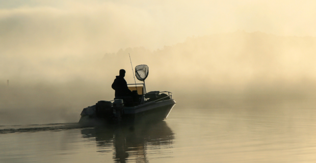 Silhouette of a fisherman in his boat with his fishing gear, early in the morning on a fog covered lake. 