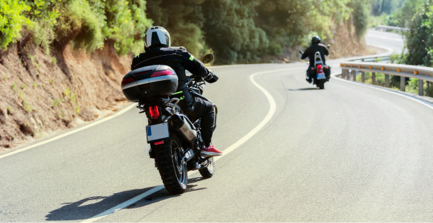 Bikers wearing motorcycle gear driving along a curvy road that’s on a hillside with forest to the left of the riders. 