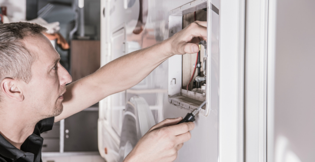 RV Repair Technician in his 40s looking for potential problem inside Travel Trailer propane gas heating system compartment.