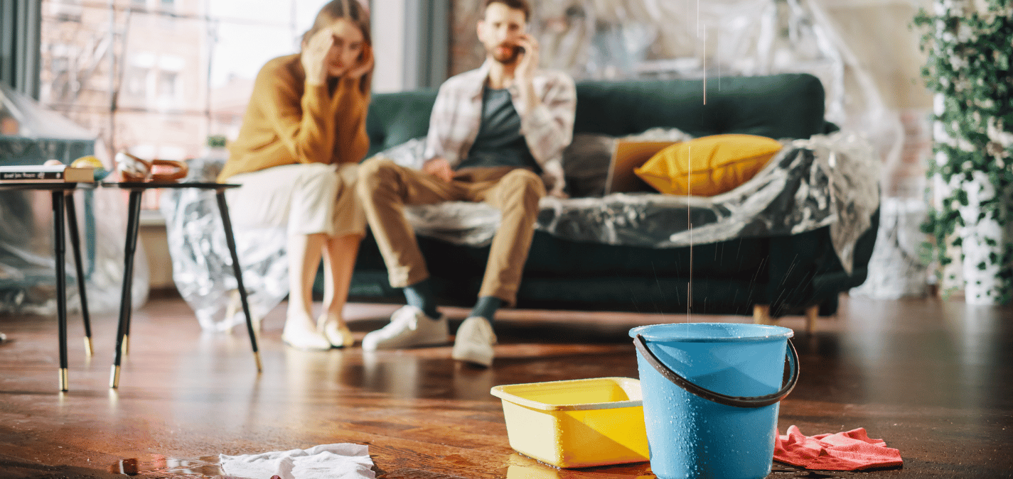 Water damage in a living room with buckets on the floor and furniture covered in plastic to catch leaks.