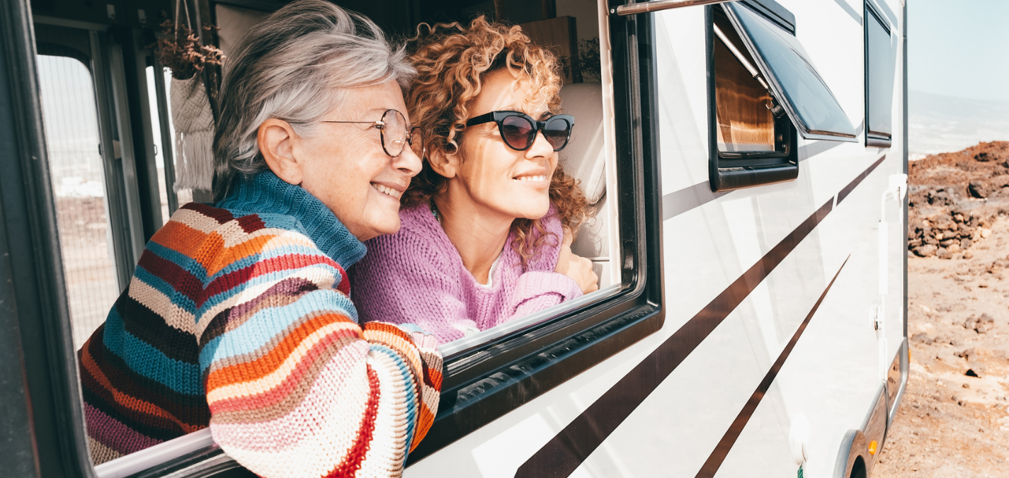 Two people lean out of the window of an RV parked in a rocky outdoor setting. They are wearing colorful sweaters and appear to be enjoying the scenic view.