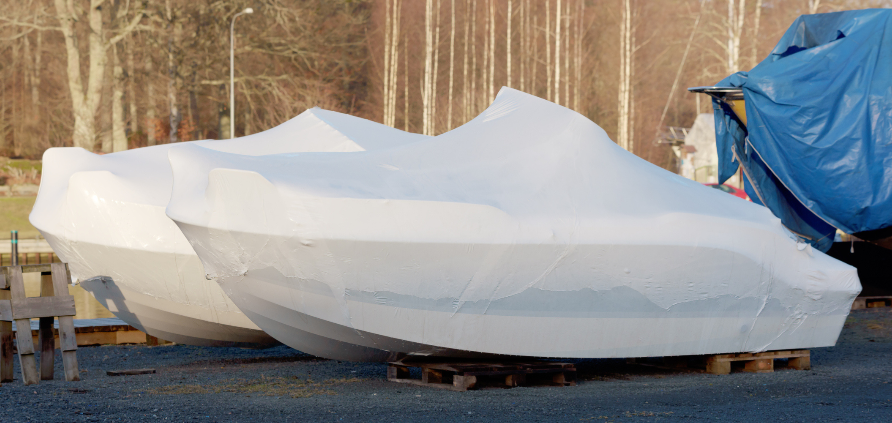 Two boats covered in white shrink wrap for winter storage, placed on wooden pallets outdoors near trees and a blue tarp-covered structure..