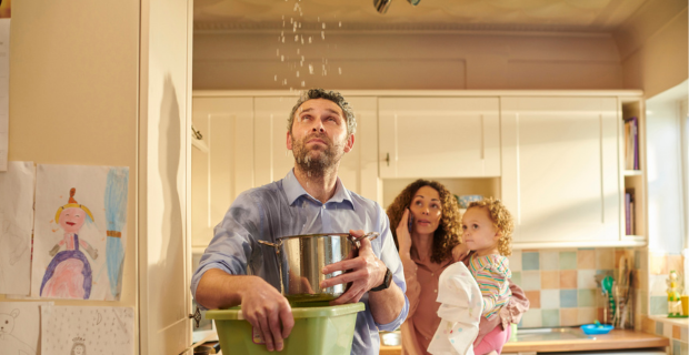 A man stands with a pot, catching water that is pouring from his kitchen ceiling. In the background, his partner is speaking on the phone, with a toddler on her hip. 
