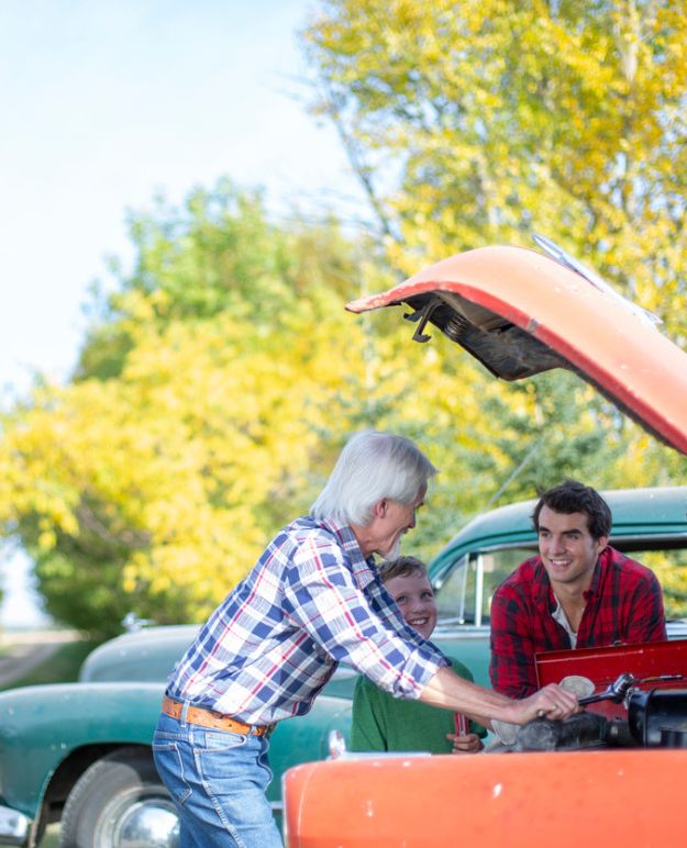 A grandparent works under the hood of a classic car while their son and grandson watch.