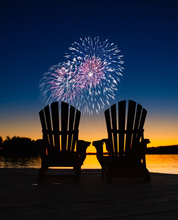 Fireworks over a still lake as the evening turns to dusk. Treescape silhouettes the lake with two Adirondack chairs in the foreground.
