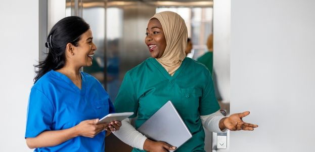 Two women in hospital scrubs, smiling and talking as they leave the elevator at their hospital. 