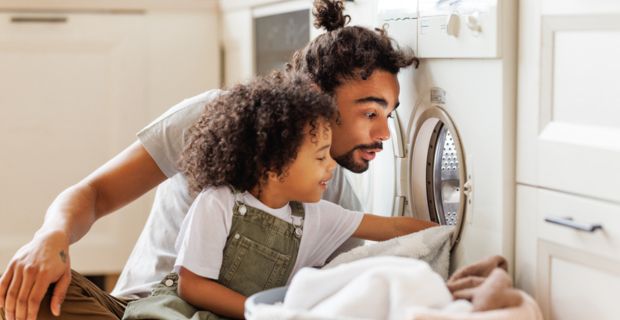A father unloading clothed from the dryer with the help of his toddler. 