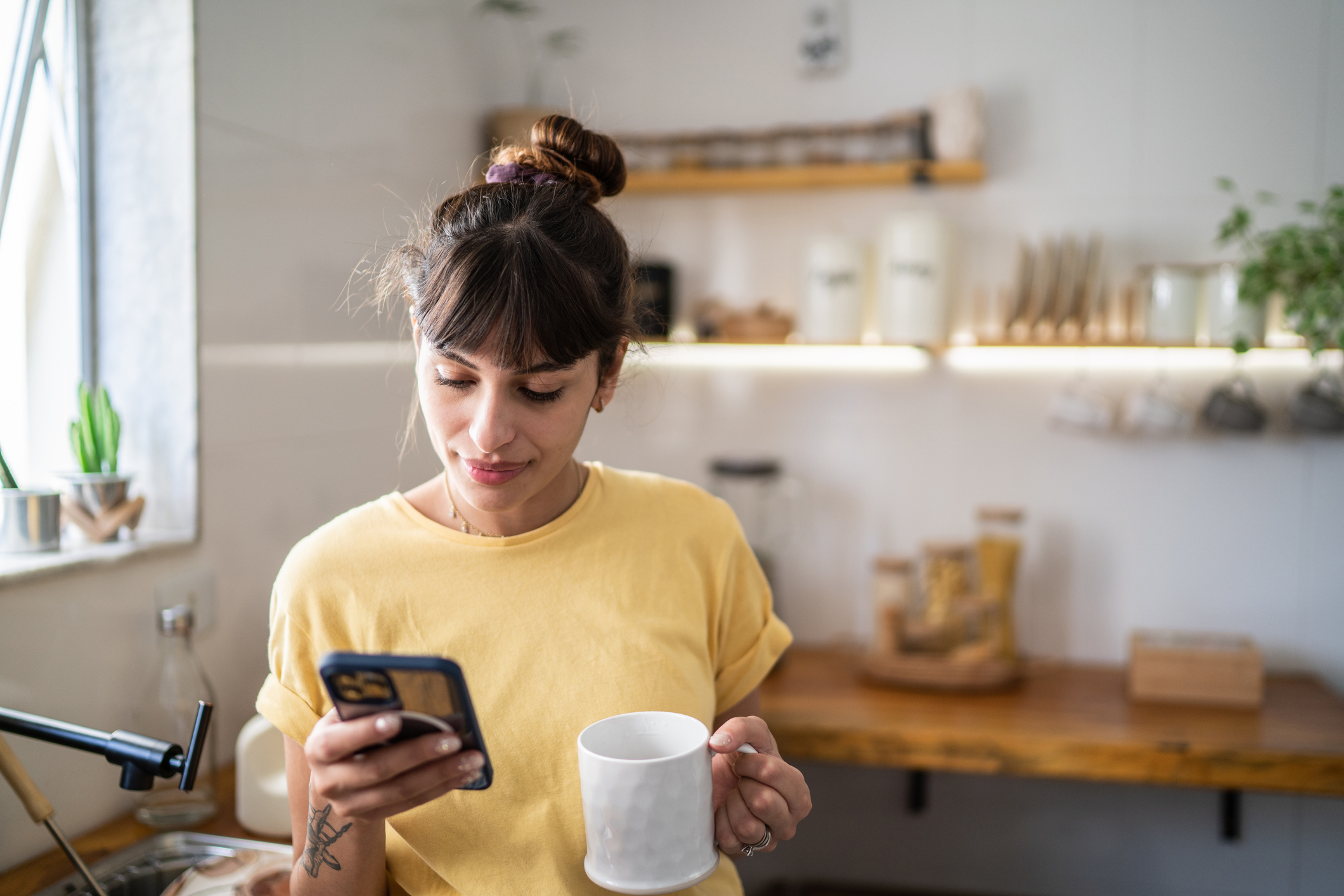 Young woman using phone while drinking from mug at home 