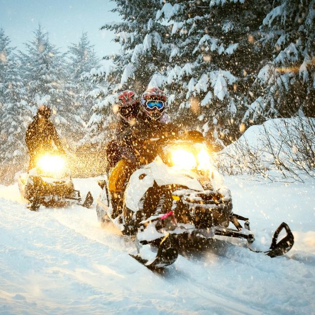 A group of riders with their headlights on, snowmobiling on a snowy day.