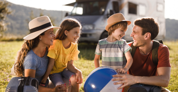 A family of four sits on the grass in front of a motorhome, dressed in casual summer clothes; one child is holding a beach ball.