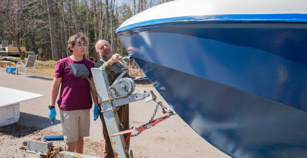 A father and his teen-aged son painting an old boat they are restoring in the driveway of their home.