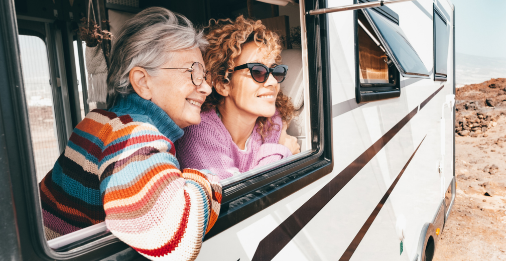 Two people lean out of the window of an RV parked in a rocky outdoor setting. They are wearing colorful sweaters and appear to be enjoying the scenic view.