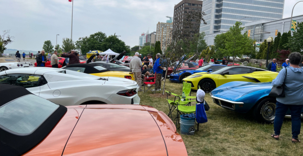 An image of a variety of both classic and modern collector cars. There are people standing around the cars admiring the beauty of the collection! In the background medium height business buildings can be seen