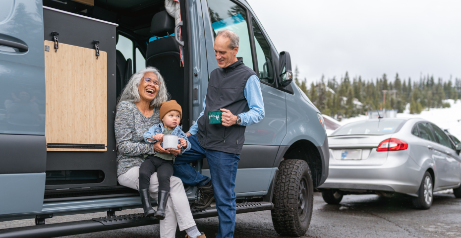 A toddler sitting on his grandparents lap in the doorway their motorhome while parked at a skiing resort.