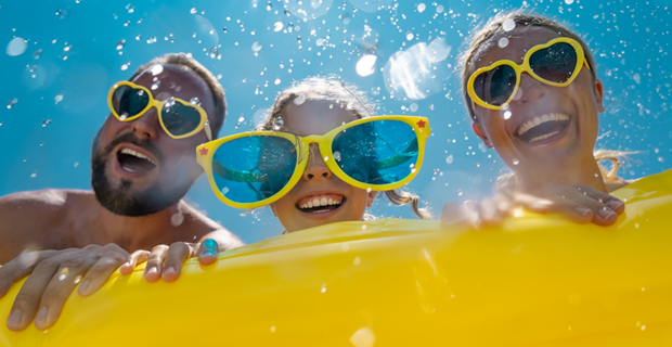Fun summer scene with three people behind a yellow pool float, wearing colorful sunglasses and goggles, with water splashes and a bright blue sky.