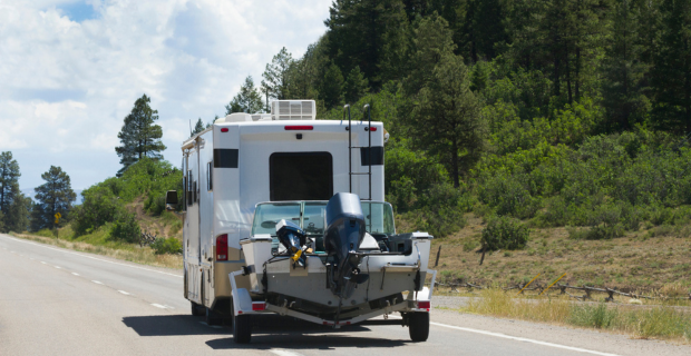 A Motorhome is towing a boat down an open road in the summer.