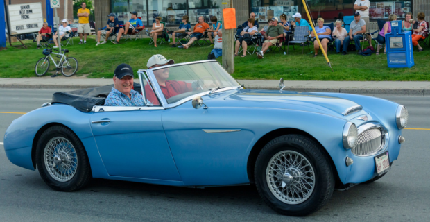 Driver & passenger wearing ball caps cruise in a 1960s Austin Healey 3000 Saturday evening on Mountain Road during 2015 Atlantic Nationals Automotive Extravaganza.