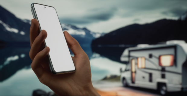  A hand holding a smartphone with a blank white screen. There is an RV parked by a lake with mountains in the background. 