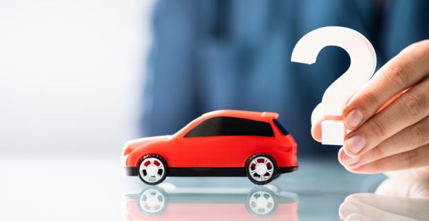 A red toy car set ontop of a table with a person holding up a question mark behind the toy car.