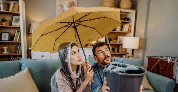 A man sitting next to his partner on their couch in their home holding a bucket to catch water leaking from the roof while his partner is holding an umbrella.