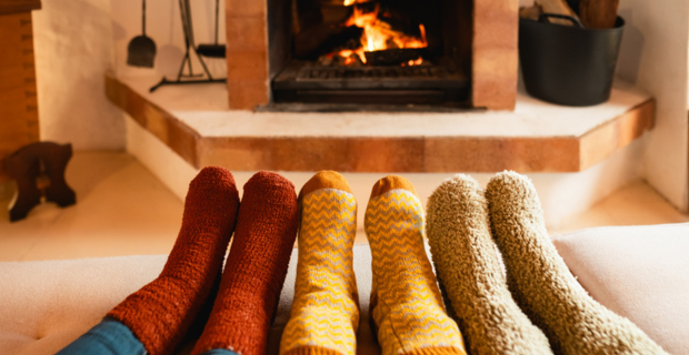 Three sets of feet all wearing different coloured socks in front of a fire staying warm in the winter. 