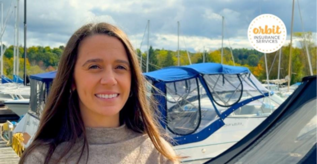 A person with long hair stands on a dock talking about marine insurance. They’re smiling and wearing a beige sweater while several boats are docked behind them. 