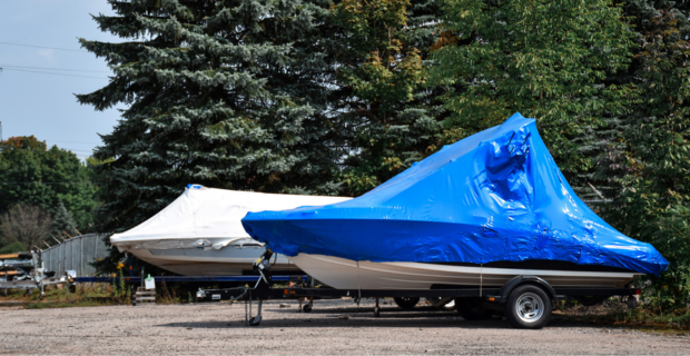 Two boats sitting in a winter boat storage lot next to ever green trees. One has a white cover and the other has a blue cover. 