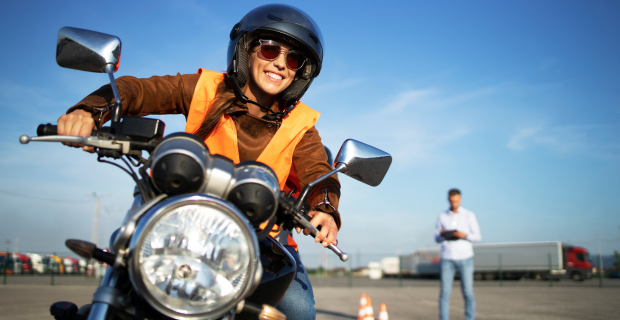 A young woman is seen driving a motorcycle during a motorcycle assessment on a sunny day.