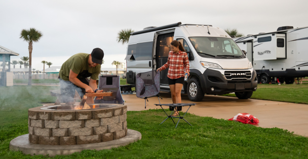 A man and women standing near a fire pit. There is an RV in the background.