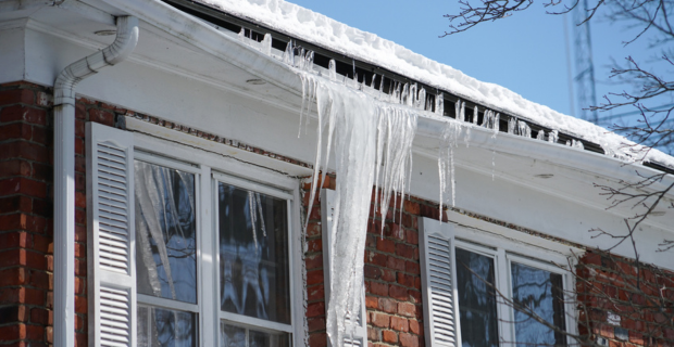 Icicles hanging from the edge of a snow-covered roof on a brick house with white shutters, under a bright blue winter sky.  