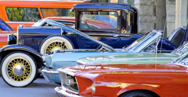 A collection of stunning vintage, collector, and classic cars are proudly on display under the summer sun. The gorgeous orange, blue, and green cars are displayed with the hood up or down.