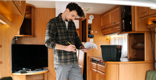 An image of a man standing in the centre of his RV. The man has dark stylish hair with a goatee. He is holding a clipboard with paper and is using a pencil to check off items from the list. On the counter next to him is a large black toolbox.
